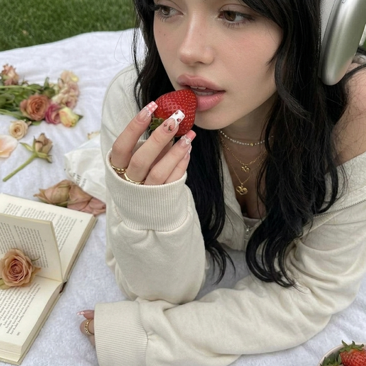 Lifestyle shot of a model wearing Berry Gingham press-on nails by MYOB Nails, featuring long square French tips with red gingham patterns, holding a fresh strawberry during an outdoor picnic.Lifestyle shot of a model wearing Berry Gingham press-on nails by MYOB Nails, featuring long square French tips with red gingham patterns, holding a fresh strawberry during an outdoor picnic.