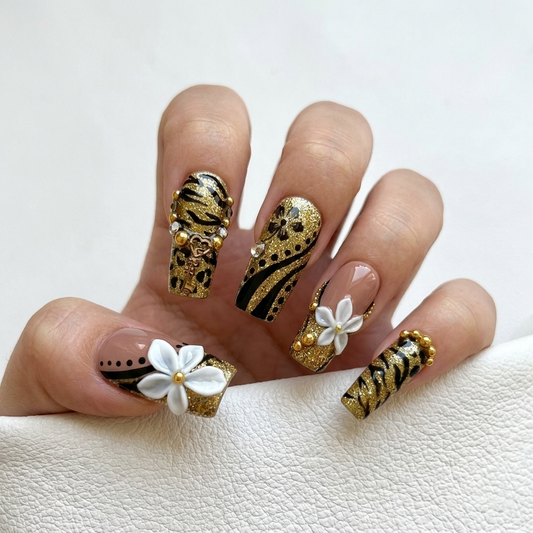 Close-up shot of MYOB Nails Millennium Disco press-on nails featuring gold glitter, black zebra print, 3D white flowers, and gold bead accents on a hand model.