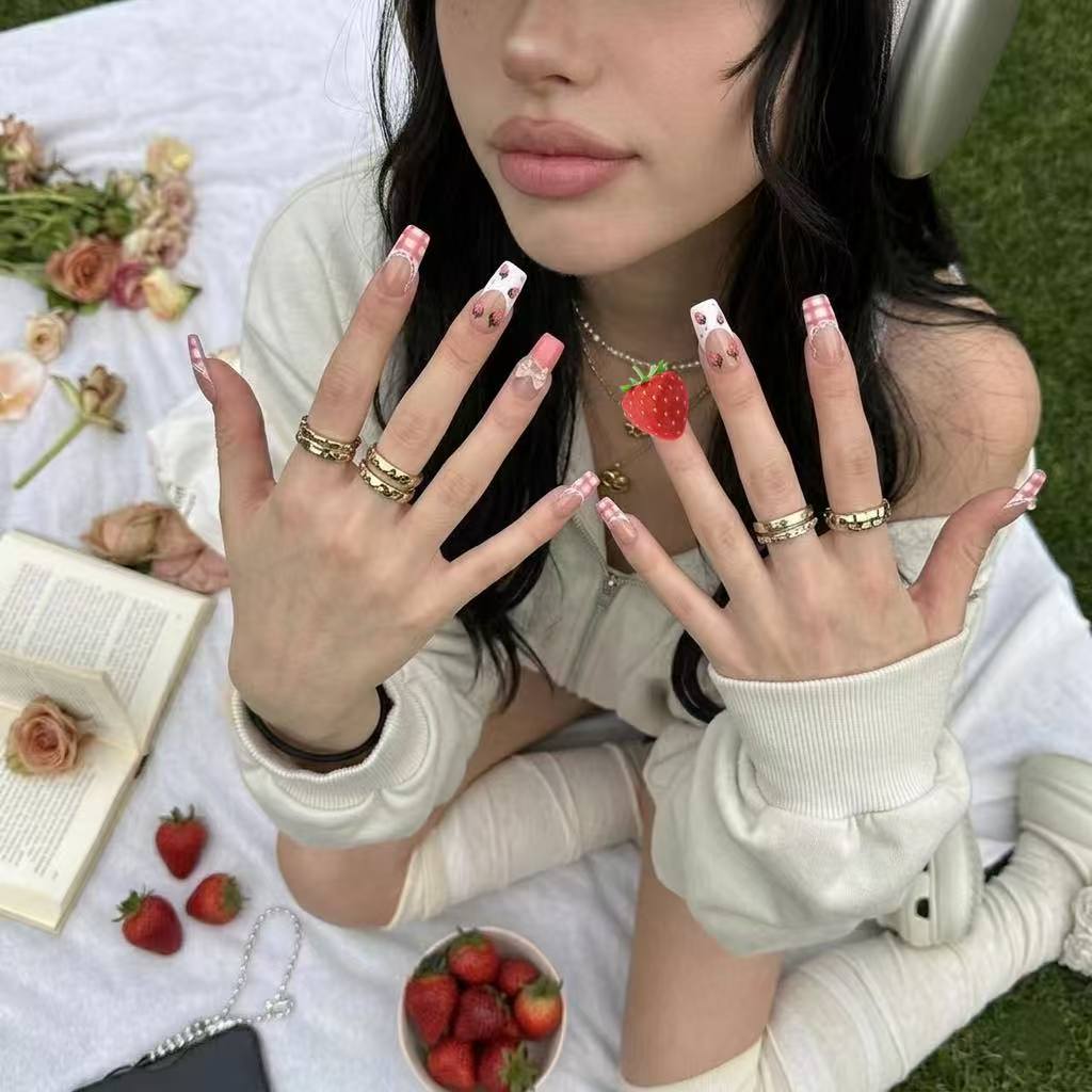 Detailed close-up shot of Berry Gingham press-on nails by MYOB Nails, showcasing long square French tips with red gingham, 3D heart motifs, and delicate bows, with the model holding a strawberry ring amidst other strawberries.
