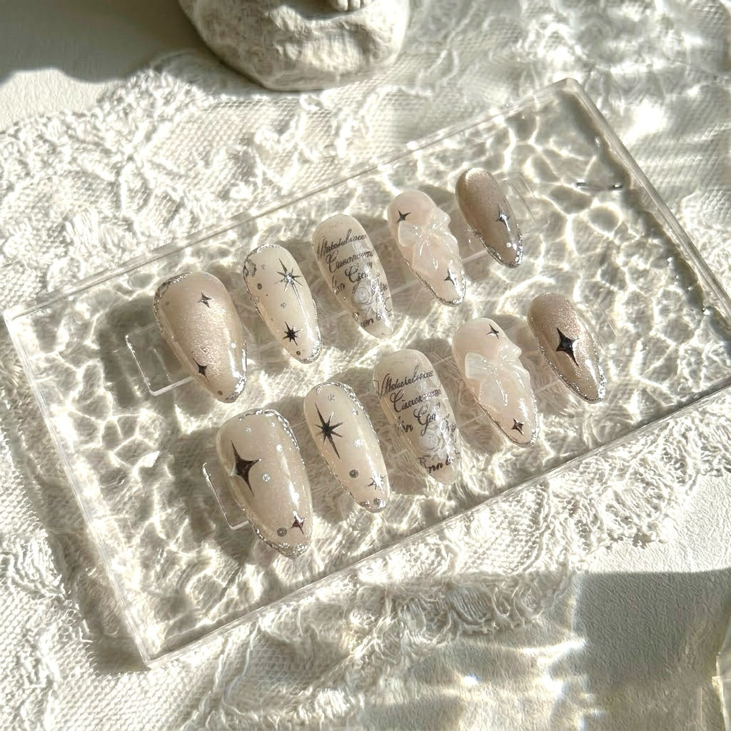 Galactic Poetry press-on nails by MYOB Nails, featuring shimmering beige cat-eye bases, silver starburst accents, and elegant script details.