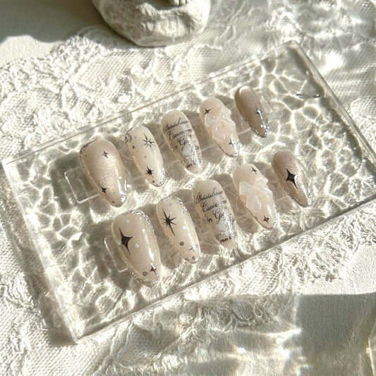 Galactic Poetry press-on nails by MYOB Nails, featuring shimmering beige cat-eye bases, silver starburst accents, and elegant script details.