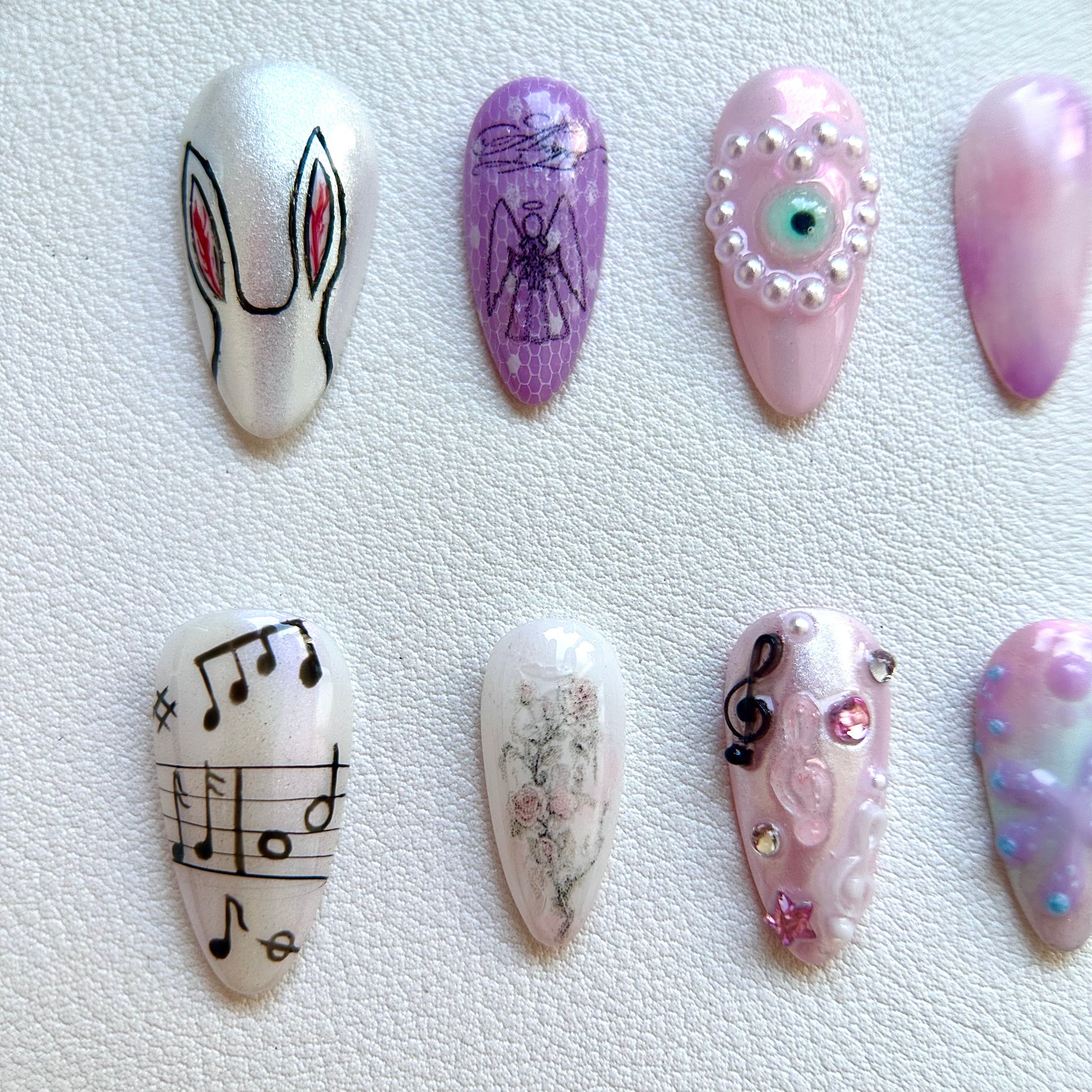 Close-up of MYOB Nails' handcrafted details featuring a minimalist hand-painted rabbit, a vintage angel sketch on purple lace, and a pink pearl-encrusted 3D evil eye charm.