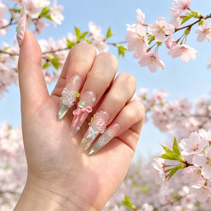 MYOB Nails A Letter from Spring press-on nails featuring clear and pink gradient almond shapes with 3D pink bows, delicate white lace patterns, and floral accents, posed against a blooming cherry blossom tree.