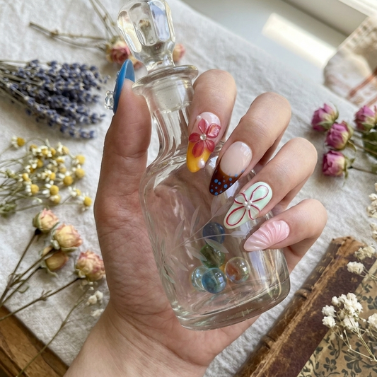 MYOB Nails Ethereal Bloom almond-shaped press-on nails worn on hand gripping a vintage glass decanter, cozy lifestyle shot surrounded by assorted fresh and dried spring florals alongside antique books, showcasing the intricate handcrafted 3D floral nail artwork, soft pastel hues and delicate glossy nail accents