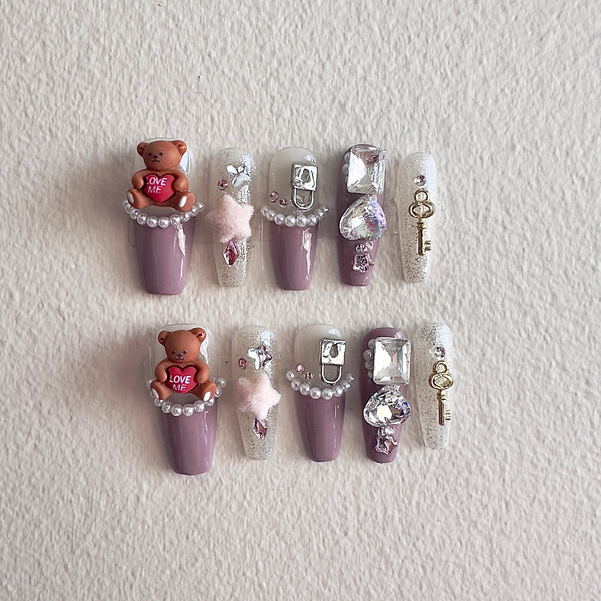 MYOB Nails elegant purple press-on nails featuring 3D brown bear charms, pearl borders, sparkling crystals, and vintage gold key accessories.