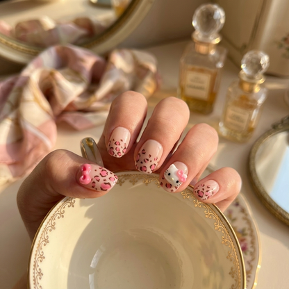 Close-up of MYOB Nails Wild Kitty press-on nails with pink leopard spots and a cute 3D cartoon cat charm, held against a vintage gold-rimmed tea cup.